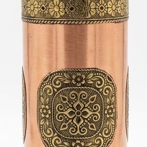 Könnte beinhalten: Eine zylindrische Wasserflasche aus Kupfer mit metallischem Glanz und passendem Schraubverschluss. Die Flasche ist mit zwei horizontalen Bändern aus geprägten Blumenmustern und einem zentralen quadratischen Feld mit filigranen Verzierungen versehen.