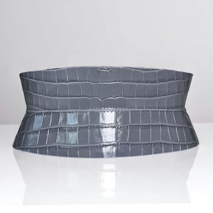 May include: A gray, wide belt with a crocodile skin texture. The belt is made of a shiny material and has a curved shape, wider in the middle and tapering towards the ends. The belt is designed to be worn around the waist.