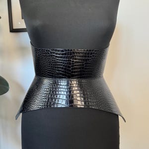 May include: A black, wide corset belt with a crocodile skin texture. The belt is displayed on a black mannequin. The belt has a wide, structured design that cinches at the waist and flares out at the hips.