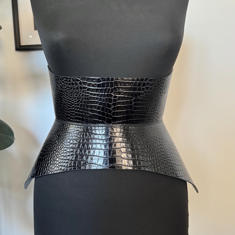 May include: A black, wide corset belt with a crocodile skin texture. The belt is displayed on a black mannequin. The belt has a wide, structured design that cinches at the waist and flares out at the hips.