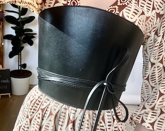 May include: A black leather corset belt with a wide band and long ties. The belt is tied in a bow at the front and has a smooth, shiny finish.