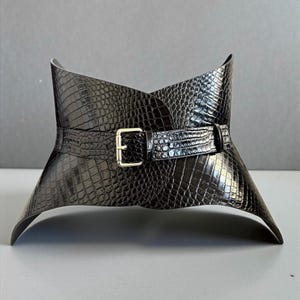 May include: A black, wide corset belt with a textured, crocodile-like pattern. The belt features a gold-colored buckle and a wrap-around design. The belt is a fashion accessory.