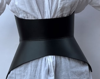 Könnte beinhalten: Ein schwarzes Lederkorsett mit spitzem, ausgestelltem Design. Das Korsett wird über einem weißen Hemd getragen.