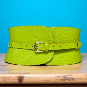 May include: A bright green, wide waist belt with a textured, crocodile-like pattern. The belt features a smaller, adjustable strap with a gold-colored buckle. The background is a solid blue.