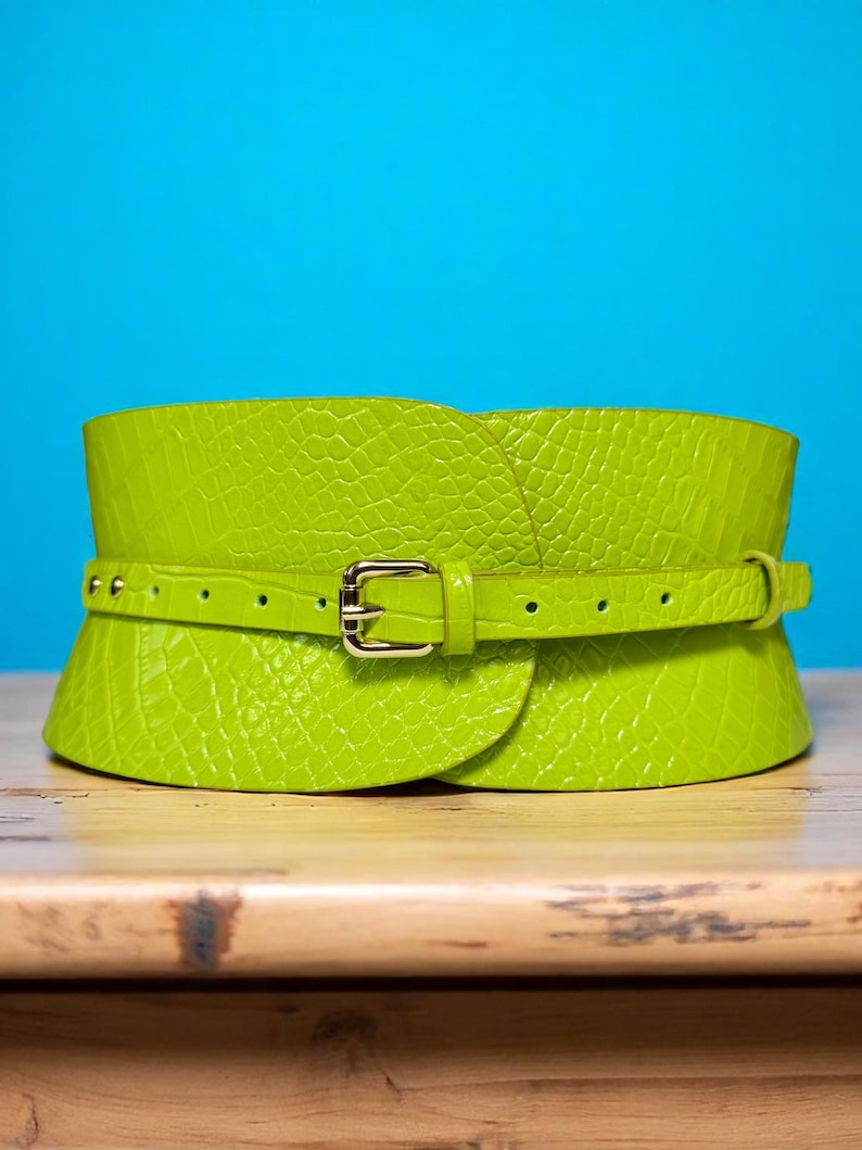 May include: A bright green, wide waist belt with a textured, crocodile-like pattern. The belt features a smaller, adjustable strap with a gold-colored buckle. The background is a solid blue.