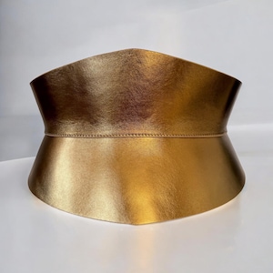 Pode incluir: Um cinto de couro dourado com uma forma larga e curva. O cinto é feito de um material liso e brilhante e tem um design simples e elegante.