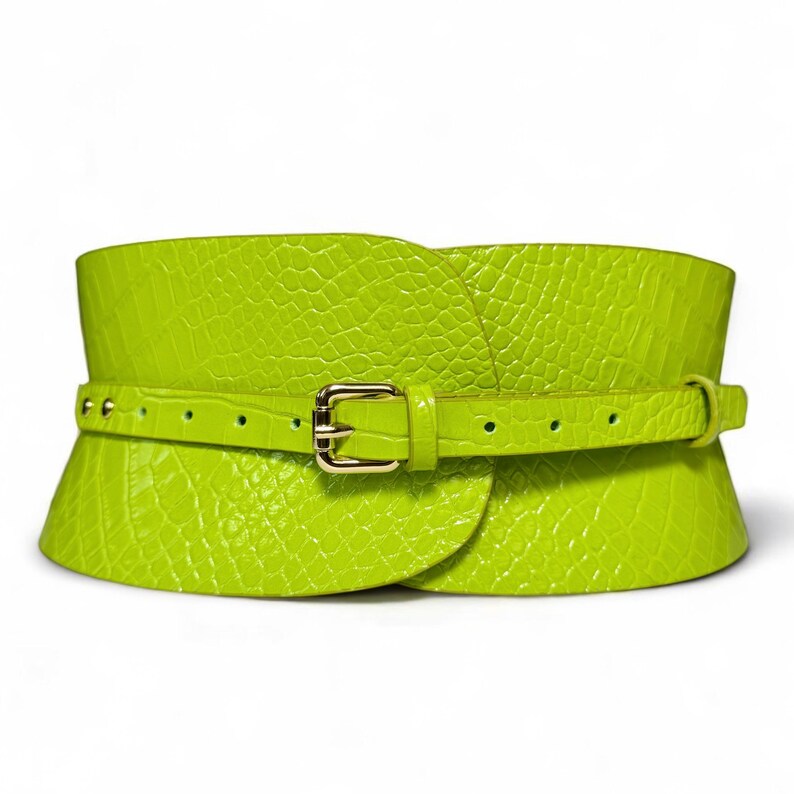 May include: A wide, lime green belt with a textured, crocodile-like pattern. The belt features a smaller, matching belt that fastens with a gold-colored buckle. The design is a wide corset style, with a wrap-around shape.
