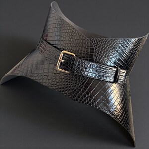 May include: A black, crocodile-textured leather collar with a gold-colored buckle. The collar has a wide, flared design and a belt closure. The overall aesthetic is sleek and modern, suitable for fashion or costume use.