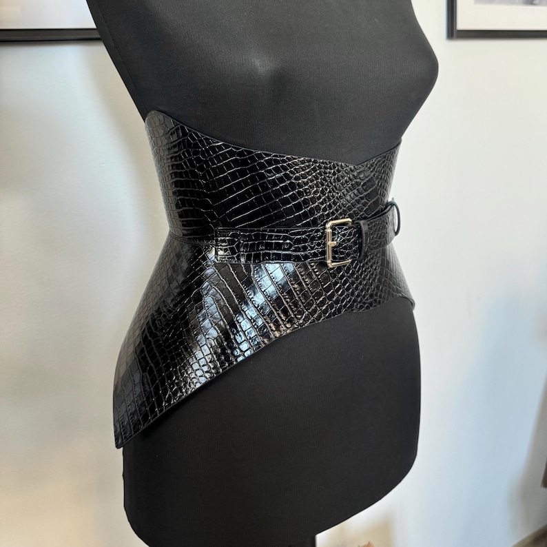 May include: A black, wide waist belt with a crocodile texture. The belt is made of a shiny material and features a silver-colored buckle. The belt is displayed on a black mannequin.