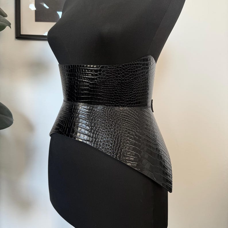 May include: A black, wide waist belt with a crocodile texture is displayed on a black mannequin. The belt has a structured design with a wider central panel and a flared lower edge. The belt is designed to cinch the waist.