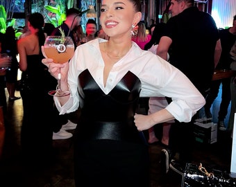 May include: A woman wearing a white button-down shirt over a black leather corset and a black skirt. She is holding a cocktail glass with a pink drink and a sprig of rosemary.
