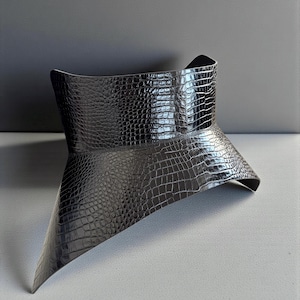 May include: A black, textured, faux-leather collar with a crocodile skin pattern. The collar has a wide, flared design that tapers to a point in the front. The collar is a fashion accessory.