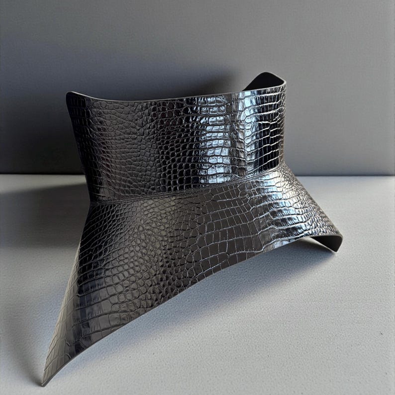 May include: A black, textured, faux-leather collar with a crocodile skin pattern. The collar has a wide, flared design that tapers to a point in the front. The collar is a fashion accessory.