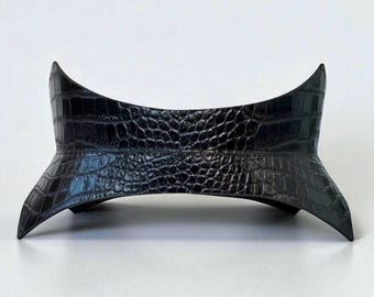 Pode incluir: Um cinto de couro preto com textura de crocodilo e um design curvo. O cinto tem um centro largo e afunila em pontas em cada extremidade. O cinto é provavelmente projetado para ser usado na cintura.