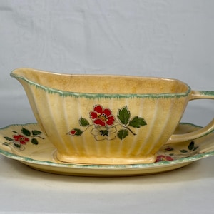 May include: A vintage yellow gravy boat and saucer set. The set features a ribbed design with red floral accents and green trim. The gravy boat has a handle and spout, and the saucer is scalloped.