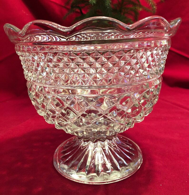 May include: A clear glass bowl with a scalloped rim and a diamond-shaped pattern. The bowl sits on a pedestal base with a fluted design.