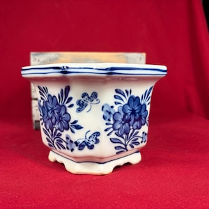May include: A white ceramic planter with a hexagonal shape, featuring blue floral and butterfly patterns. The rim and base are accented with thin blue lines, and the design includes clusters of flowers and small butterflies on the sides.