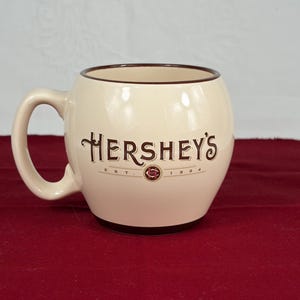 May include: Cream-colored Hershey's mug with a rounded shape and a brown rim and base. The Hershey's logo is prominently displayed in brown script. The mug has a large handle and is set against a red background.