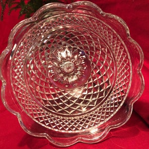 May include: A clear glass cake stand with a scalloped edge and a diamond pattern. The stand is designed for serving desserts or other treats.