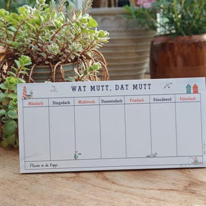 Könnte beinhalten: Ein weißer Wochenplaner mit dem Text "WAT MUTT, DAT MUTT" und Wochentagen auf Deutsch. Der Planer hat dekorative Illustrationen eines Leuchtturms, Vögeln und Häusern. Der Satz "Fluver in de Kopp" steht unten.