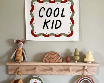 May include: A wall-mounted shelf with decorative items, including a "COOL KID" sign, a toy mouse, wooden toys, and a rainbow-shaped wall hanging. The sign has a white background with red and green trim.