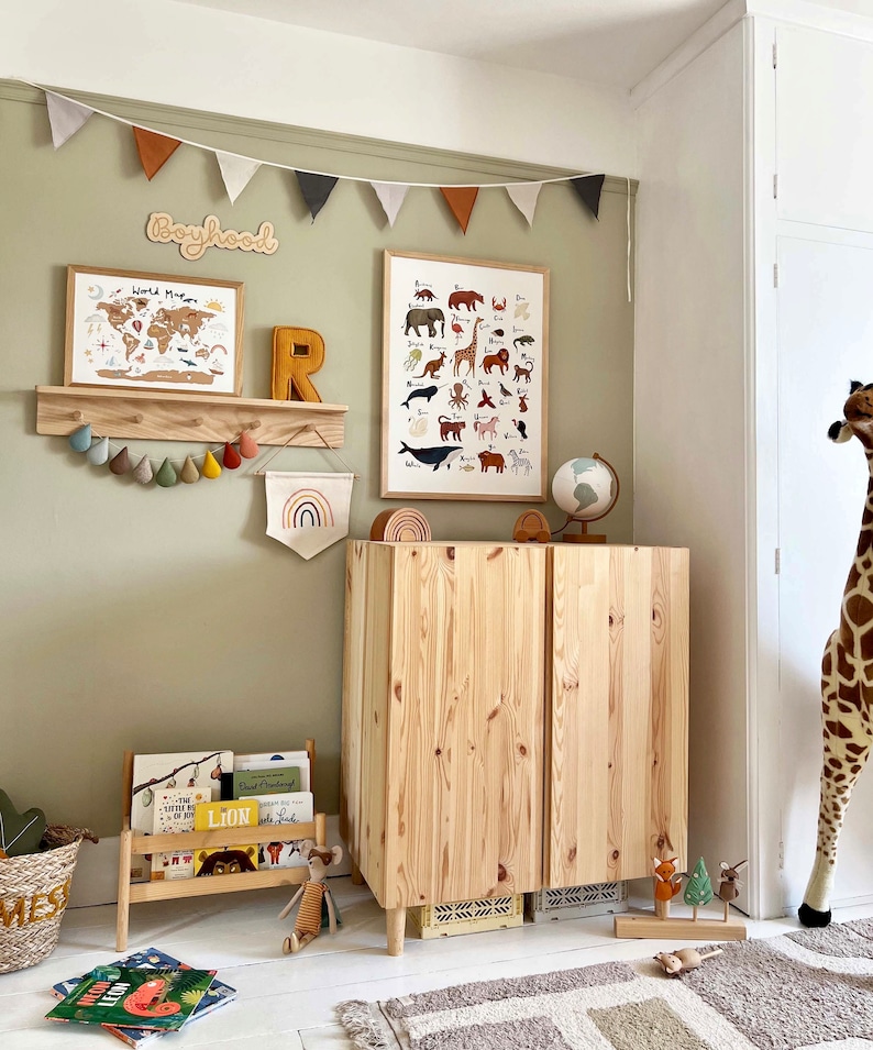 May include: A light green nursery with a wooden cabinet, a globe, a world map, and a wooden shelf with a wooden letter R and a banner of colorful felt teardrop shapes. The wall is decorated with a banner of colorful triangle flags and a wooden sign that says "Boyhood".
