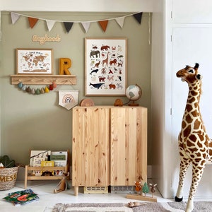 May include: A light wood cabinet with two doors, a wooden shelf with a world map print and a wooden letter R on top. There is a stuffed giraffe standing next to the cabinet. The wall behind the cabinet is painted a light green color and has a string of flags hanging from it. The flags are in various colors, including red, orange, and brown. The text "Boyhood" is written on the wall in a wooden font.