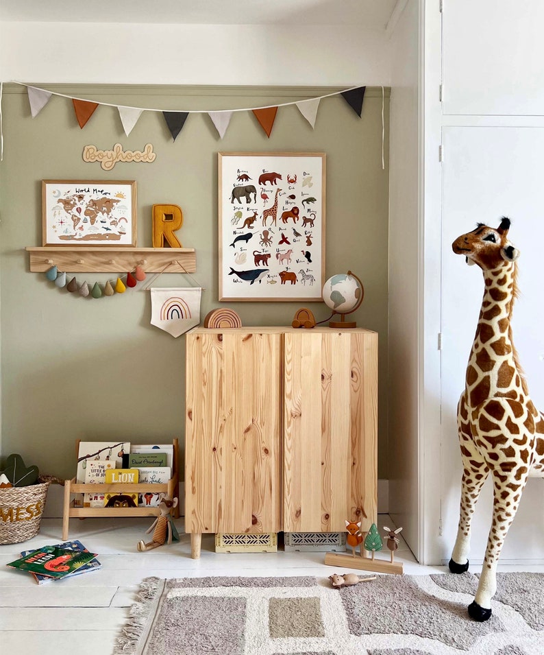 May include: A light wood cabinet with two doors, a wooden shelf with a world map print and a wooden letter R on top. There is a stuffed giraffe standing next to the cabinet. The wall behind the cabinet is painted a light green color and has a string of flags hanging from it. The flags are in various colors, including red, orange, and brown. The text "Boyhood" is written on the wall in a wooden font.