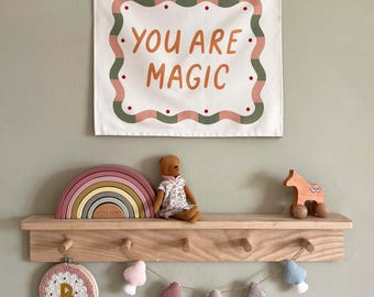 May include: A wooden shelf displays a rainbow-shaped toy, a teddy bear, and a wooden horse toy. Above, a wall hanging with the text "YOU ARE MAGIC" is framed by a pink and green border. Below, a garland of felt mushrooms and a framed embroidery piece.