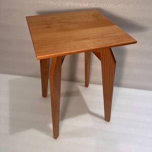 K&ouml;nnte beinhalten: Kleiner, quadratischer Beistelltisch aus Holz mit nat&uuml;rlicher Oberfl&auml;che. Die Tischplatte wird von vier abgewinkelten Beinen getragen. Der Tisch ist etwa 61 cm hoch und 46 cm breit.