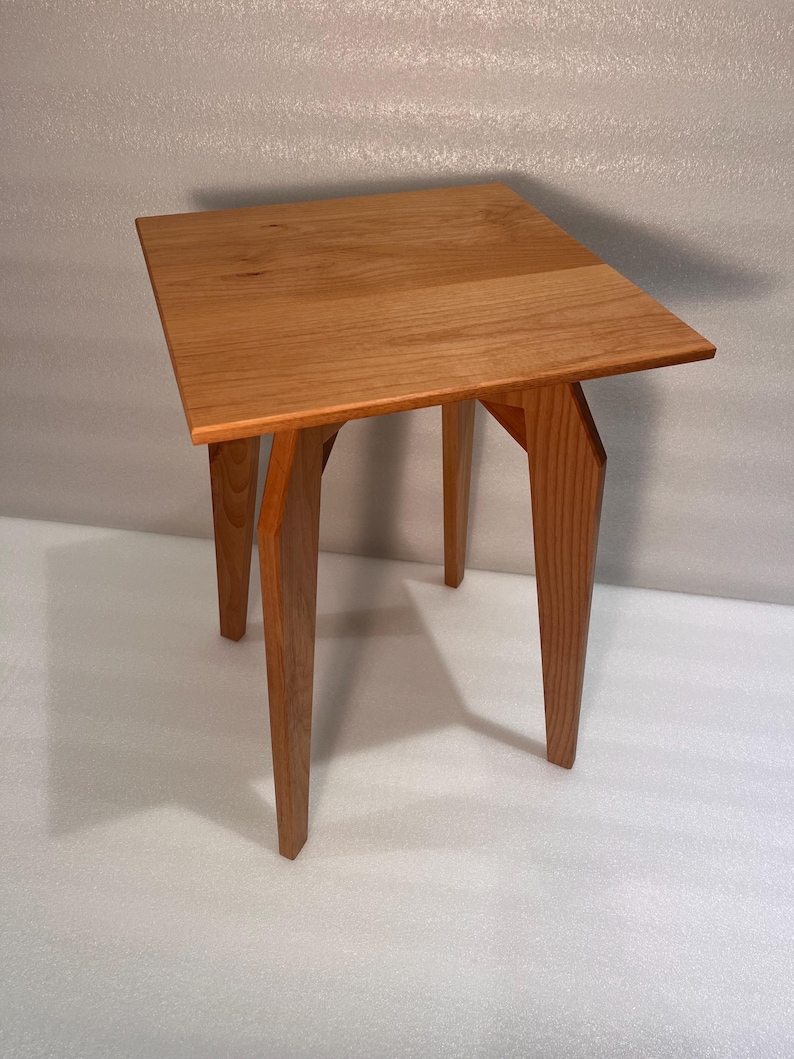 K&ouml;nnte beinhalten: Kleiner, quadratischer Beistelltisch aus Holz mit nat&uuml;rlicher Oberfl&auml;che. Die Tischplatte wird von vier abgewinkelten Beinen getragen. Der Tisch ist etwa 61 cm hoch und 46 cm breit.