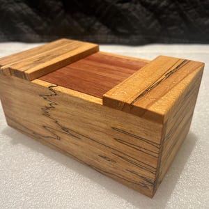 May include: A rectangular wooden box featuring a light tan exterior with dark, jagged grain patterns and a smooth, reddish-brown center panel on the lid. The box has a flat top with two raised wooden sections on either side of the central panel.