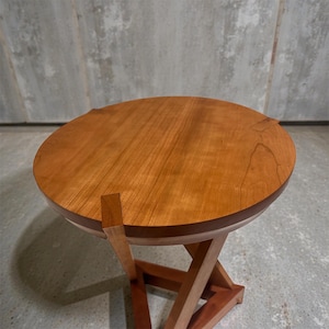 May include: A handcrafted wooden side table with a round top and a unique, geometric base. The tabletop is a warm, reddish-brown color, and the base is constructed with lighter-toned wood. The table is designed for indoor use and is a functional piece of furniture.