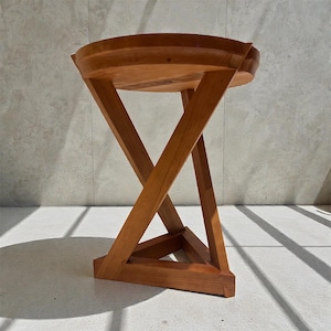 May include: A wooden side table with a round top and a unique, geometric base. The table is made of light brown wood and features an X-shaped support structure. The table is approximately 18 inches in diameter and 20 inches tall.