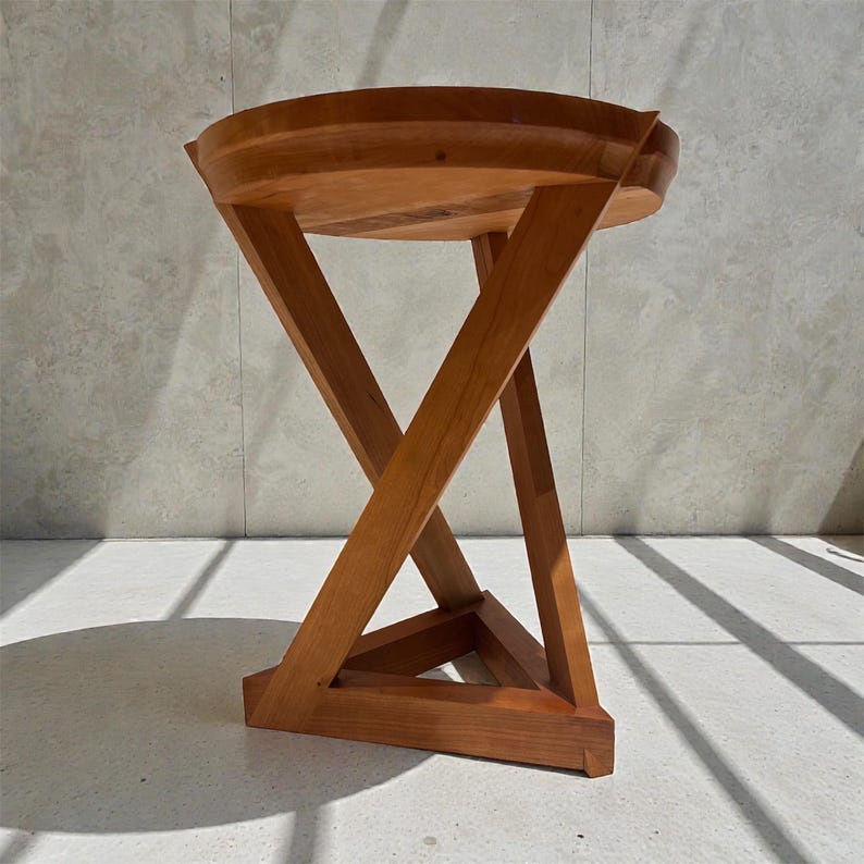 May include: A wooden side table with a round top and a unique, geometric base. The table is made of light brown wood and features an X-shaped support structure. The table is approximately 18 inches in diameter and 20 inches tall.