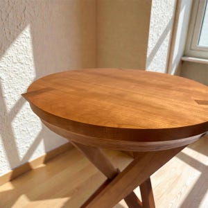 May include: A round wooden side table with a warm, reddish-brown finish. The tabletop is approximately 24 inches in diameter and rests on a crossed-leg base. The table is made of solid wood and has a smooth, polished surface.