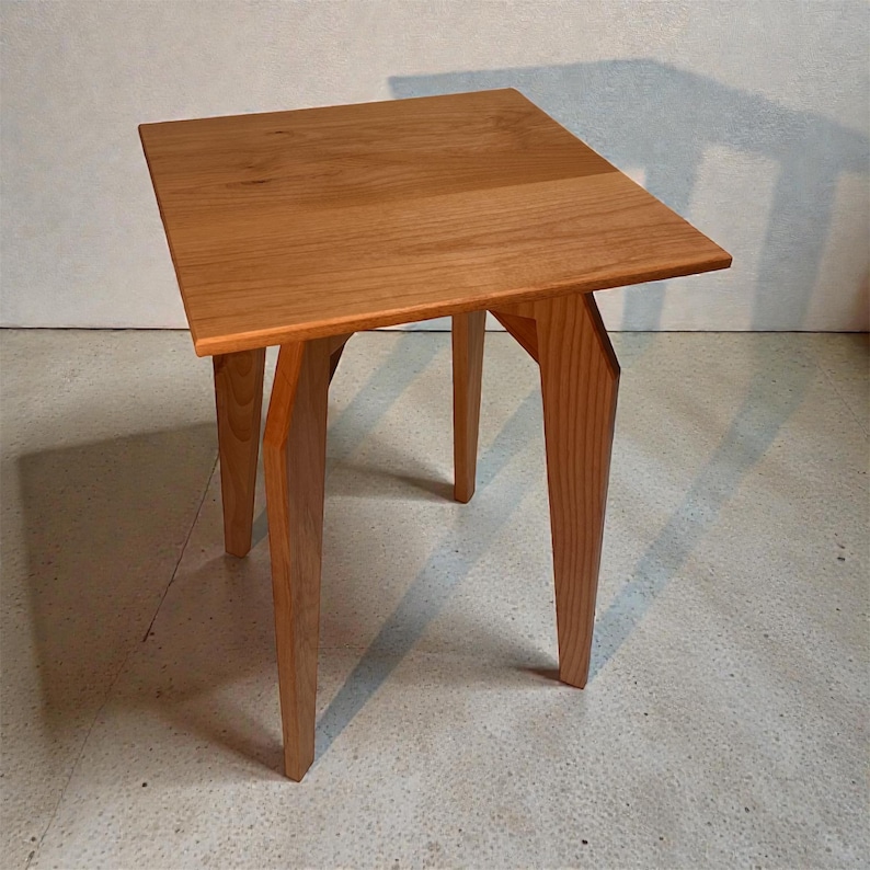 K&ouml;nnte beinhalten: Kleiner, quadratischer Beistelltisch aus Holz mit nat&uuml;rlicher Oberfl&auml;che. Der Tisch hat eine massive Platte und drei abgewinkelte Beine. Der Tisch ist etwa 51 cm hoch und 46 cm breit.