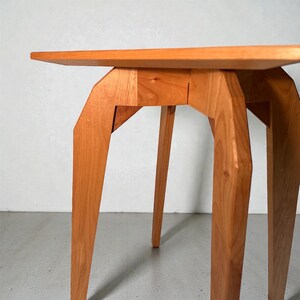 K&ouml;nnte beinhalten: Kleiner Beistelltisch aus hellem Holz. Der Tisch hat eine quadratische Platte und vier abgewinkelte Beine. Die Beine sind mit geometrischen St&uuml;tzen mit der Tischplatte verbunden. Der Tisch ist aus massivem Holz gefertigt und hat eine nat&uuml;rliche Maserung. Die H&ouml;he betr&auml;gt ca. 50 cm.