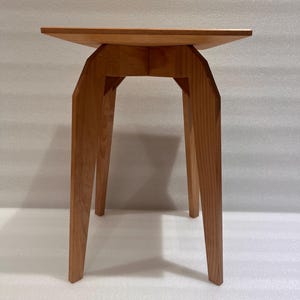 K&ouml;nnte beinhalten: Ein hellbrauner Beistelltisch aus Holz mit quadratischer Oberseite und abgewinkelten Beinen. Der Tisch ist aus Massivholz gefertigt und hat eine nat&uuml;rliche Oberfl&auml;che. Die Beine sind geometrisch geformt und verleihen einen modernen Touch. Der Tisch ist ca. 61 cm hoch.