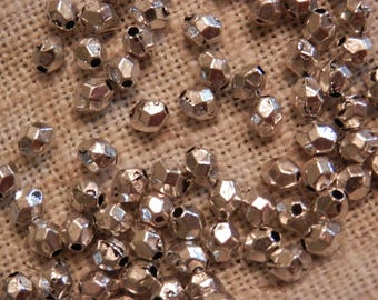May include: A close-up of a pile of silver metal beads. The beads are faceted and have a shiny finish. They are scattered on a beige fabric surface.