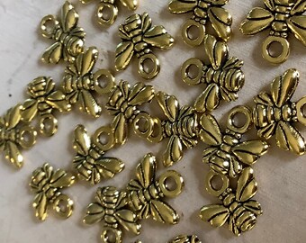 May include: A collection of small, gold-colored bee charms. Each charm features detailed wings, body segments, and a small loop for attaching to jewelry or crafts. The charms have a vintage or antique appearance.
