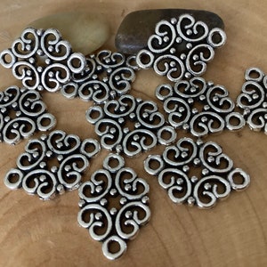 May include: Silver metal charms with a decorative, intricate design. The charms have a square shape with rounded corners and feature a repeating pattern of swirls and dots.