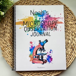 Puede incluir: Un diario encuadernado en espiral con el título "Noah's First Microscope Observation Journal". La portada presenta una ilustración en acuarela de un microscopio en rosa y azul, con salpicaduras de pintura coloridas. El diario está sobre una estera tejida.