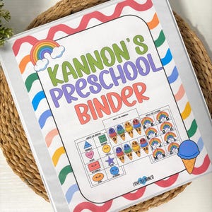 May include: A clear binder with a colorful cover featuring the text "Kannon's Preschool Binder." The cover includes a rainbow, ice cream cone, and educational matching activities for shapes, numbers, and letters. The binder is on a woven mat.