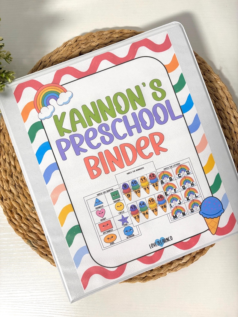 May include: A clear binder with a colorful cover featuring the text "Kannon's Preschool Binder." The cover includes a rainbow, ice cream cone, and educational matching activities for shapes, numbers, and letters. The binder is on a woven mat.