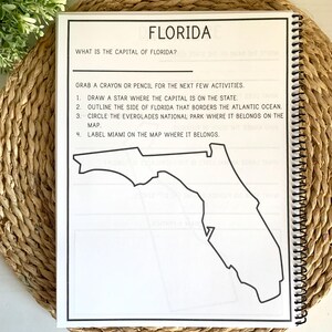 Puede incluir: Una p&aacute;gina de cuaderno con espiral titulada "FLORIDA". La p&aacute;gina incluye preguntas y actividades sobre Florida, como identificar la capital, delinear la frontera estatal y etiquetar Miami en un mapa. El cuaderno est&aacute; sobre una estera tejida.
