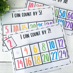 以下が含まれることがあります： 「10ずつ数えられるよ！」「5ずつ数えられるよ！」「2ずつ数えられるよ！」というタイトルの3枚のカラフルなラミネート加工された学習カード。各カードには、異なる色の数字のグリッドがあります。カードの数字は、10、20、50、60、110、120、5、10、15、20、25、2、4、6、8、10、12、14、16、18、20、22、24です。