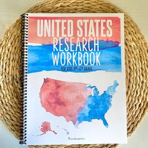 Puede incluir: Un cuaderno con espiral titulado "UNITED STATES RESEARCH WORKBOOK" para ni&ntilde;os de 3&ordm; a 6&ordm; grado. La portada presenta un mapa de EE. UU. en acuarela en rojo y azul, con un fondo de cielo en acuarela. El cuaderno est&aacute; sobre una estera tejida.