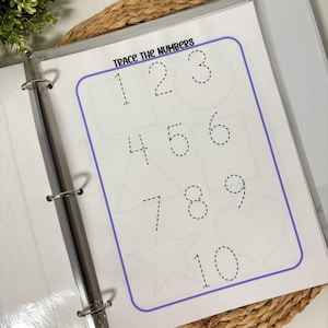 May include: A white binder open to a page with the text "Trace the Numbers." The page features numbers 1 through 10 in a dotted line format for tracing. The numbers are inside a purple border. The binder is on a woven surface.