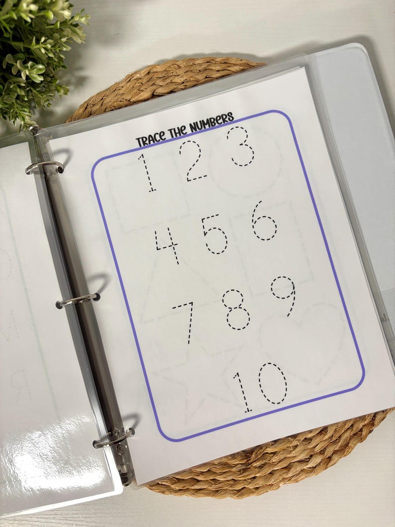 May include: A white binder open to a page with the text "Trace the Numbers." The page features numbers 1 through 10 in a dotted line format for tracing. The numbers are inside a purple border. The binder is on a woven surface.